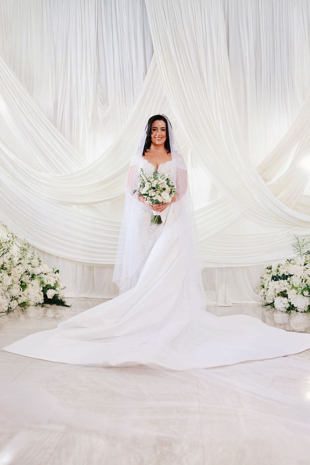 A bride in an elegant white wedding gown and cathedral veil poses against white draped fabric and floral arrangements.