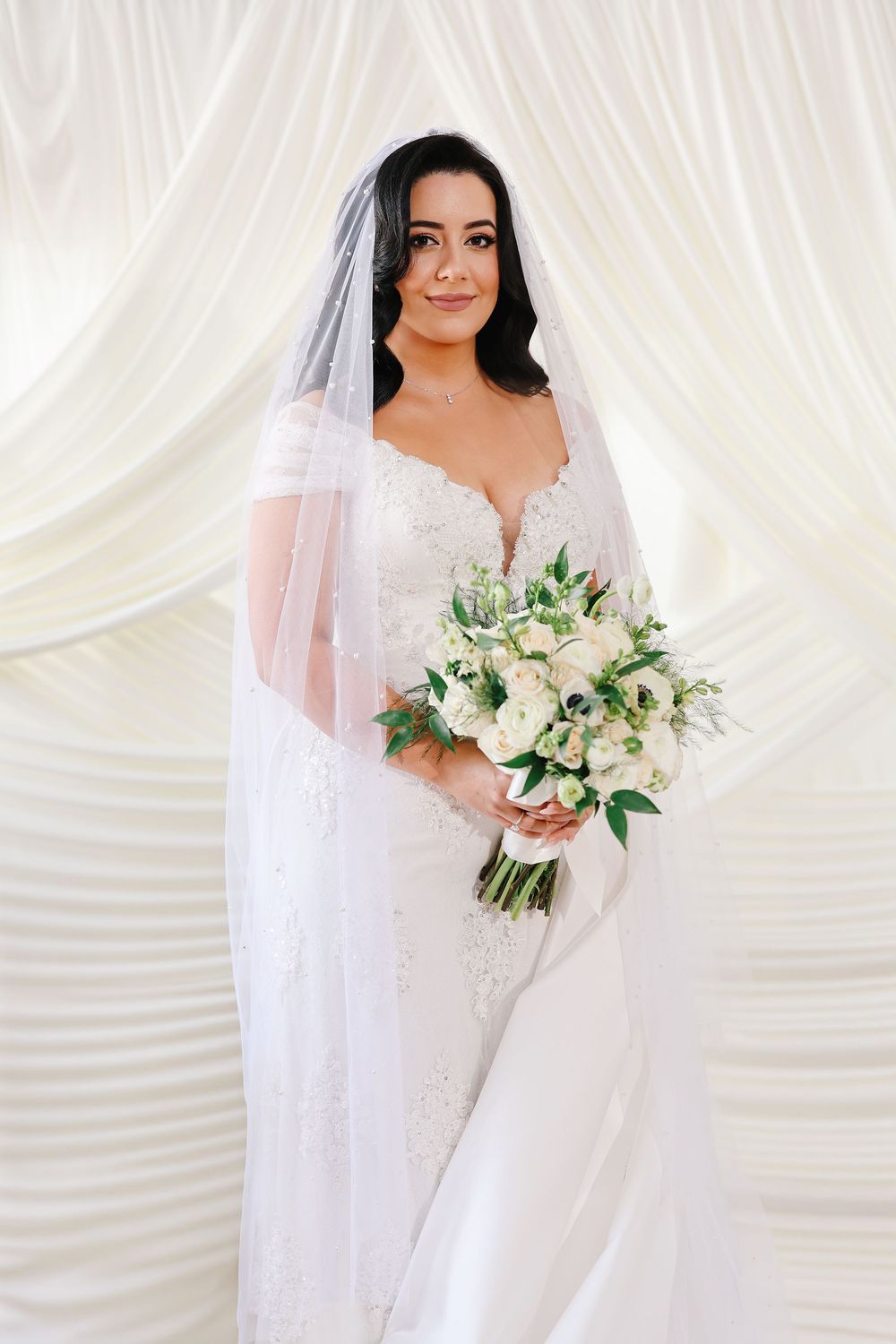 A bride in a white lace gown and long veil holds an elegant white and green floral bouquet against a draped backdrop.