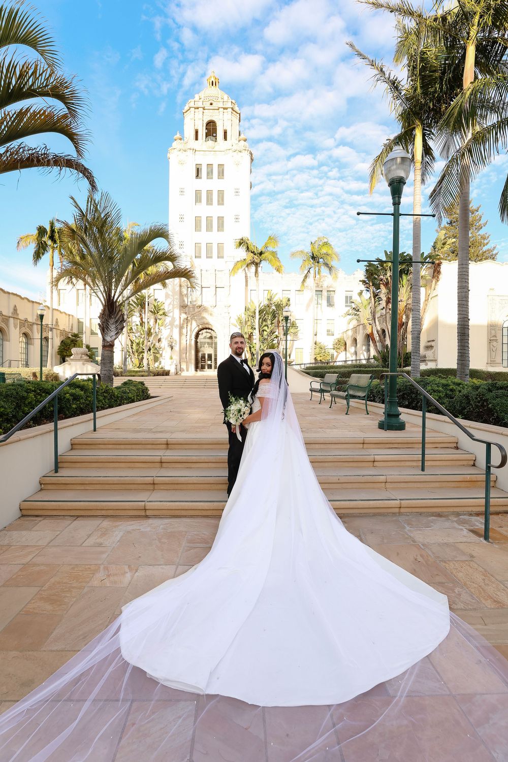 Wedding couple standing in front of a Spanish colonial church with palm trees and long trailing veil.