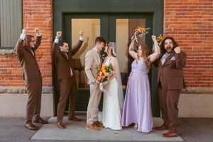 Wedding party in brown suits and purple dresses poses against historic brick building in natural light.