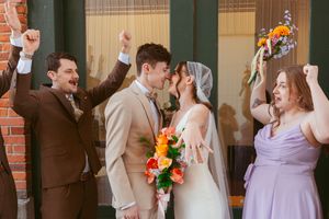 Joyful wedding party celebrates with raised arms as newlyweds share a romantic moment with sunflower bouquet.