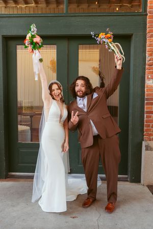 Couple in wedding attire poses playfully with celebratory gestures against a brick building.
