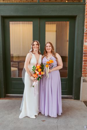 Two wedding attendees pose together holding bright floral bouquets against a brick building.