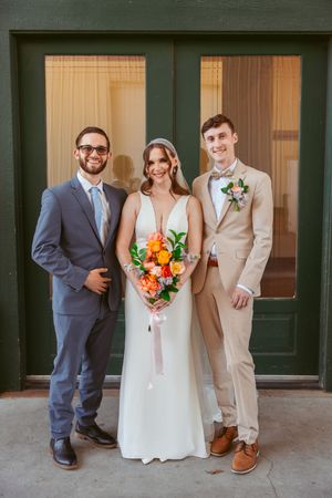 Wedding party poses for formal photos against a brick building with green doors.