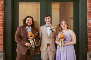 A wedding party poses casually outdoors in a mix of brown suits and light purple dresses.
