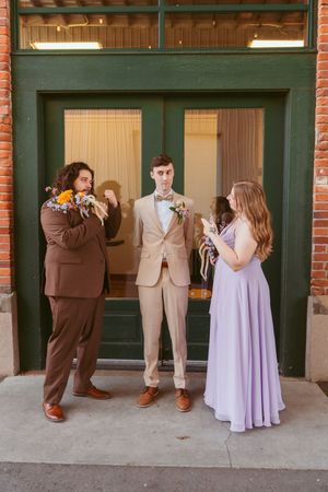 Wedding party members in brown and beige suits with lilac dress pose casually outside brick building entrance.