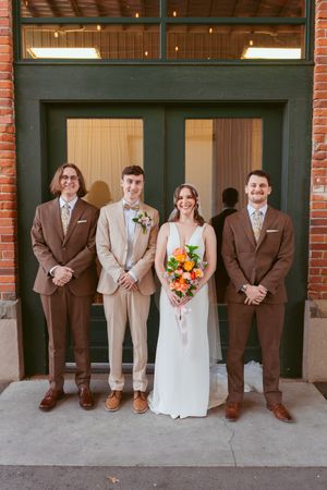 Wedding party in brown and beige suits stands with bride holding vibrant orange and yellow bouquet against brick wall.