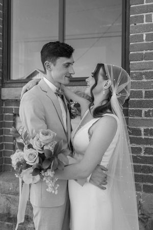 Newlywed couple in formal attire share an intimate moment against brick wall with floral bouquet.