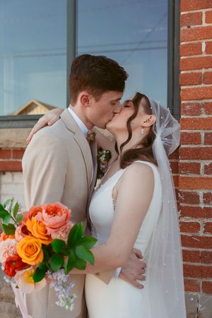 Bride and groom kiss while holding vibrant orange and pink floral bouquet against brick wall.