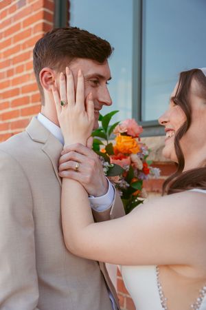 Newlywed couple shares tender first look moment against brick wall with bright bouquet in foreground.