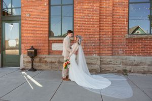 A bride in a flowing white gown with long veil stands outside a rustic brick building with stone foundation.