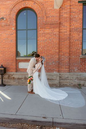 Bride in flowing veil bends back gracefully against rustic brick building with arched window.