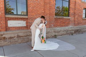 A couple shares an intimate moment against a brick building wall, with a white wedding dress trailing on the concrete sidewalk.