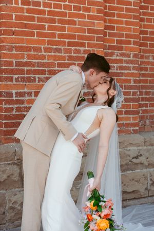 Romantic wedding couple embraces in dramatic pose against red brick wall with floral bouquet.