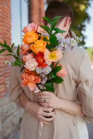 A vibrant coral and orange wedding bouquet with roses and greenery held by someone in a beige suit.