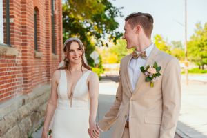 A couple in wedding attire stands together against a brick building in warm sunlight during their special day.