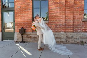 Romantic dip pose of newlyweds against brick building with flowing veil and sunlight.