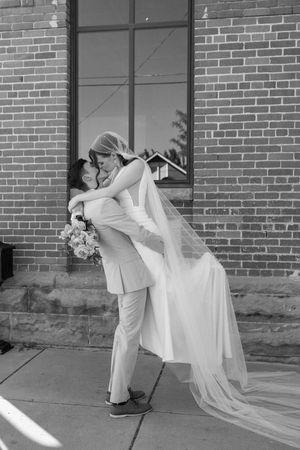 Dramatic black and white shot of groom lifting bride in flowing dress against architectural backdrop.
