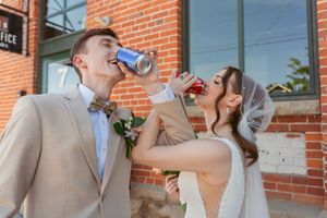 A bride and groom playfully drinking from cans in front of a brick building on their wedding day.