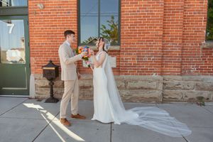 Couple in formal wedding attire pose together for photos against a rustic brick wall exterior in natural lighting.