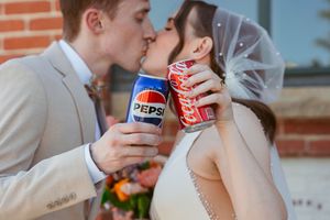 Newlyweds in wedding attire share a romantic kiss while holding Pepsi cans at a brick building.