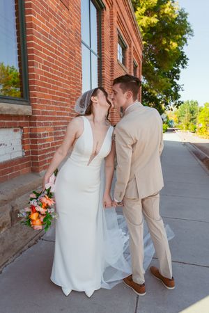 Newlywed couple in beige suit and white dress share romantic moment against brick wall exterior.