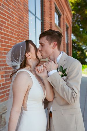 A wedding couple shares an intimate kiss against a brick wall backdrop in a romantic photography series.