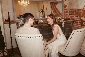 A couple sits together on a white upholstered sofa in an elegant room with exposed brick walls and chandeliers.