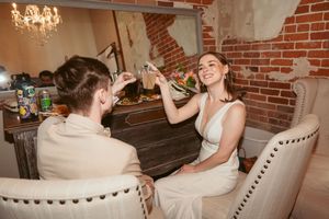 A person in a white dress sits in a chair at a rustic table while someone in beige looks on.
