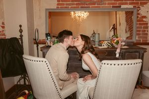 Two people sit together in elegant white studded chairs beneath a crystal chandelier in a room with exposed brick walls.