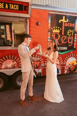 Newlywed couple shares a playful moment while eating tacos from a vibrant orange food truck at their wedding.