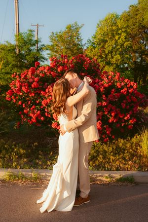 A newlywed couple embraces in a romantic sunset photoshoot with vibrant red flowers in the background.