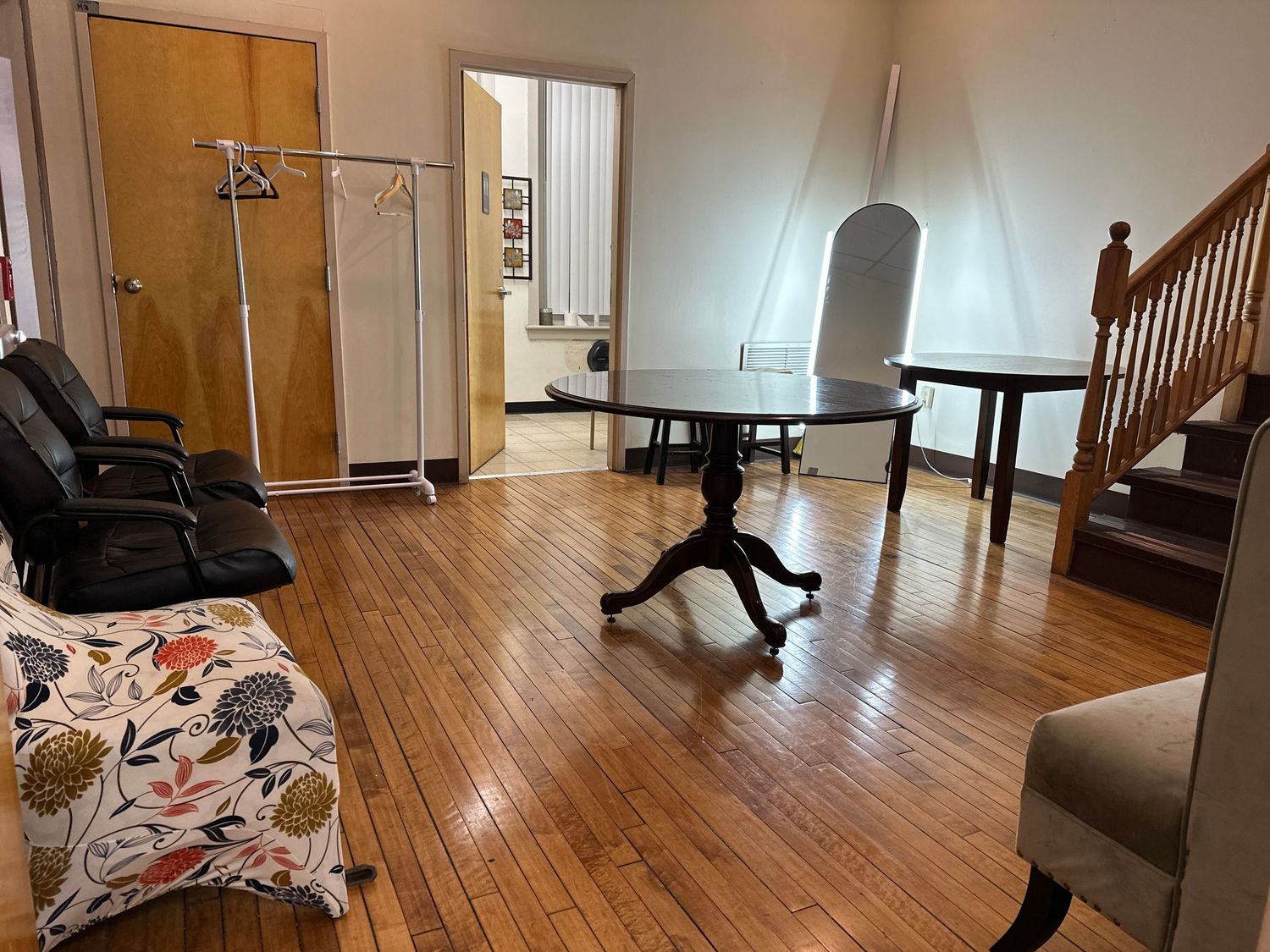 Living room interior with hardwood floors featuring a clothing rack, floral sofa, dark table and wooden staircase.