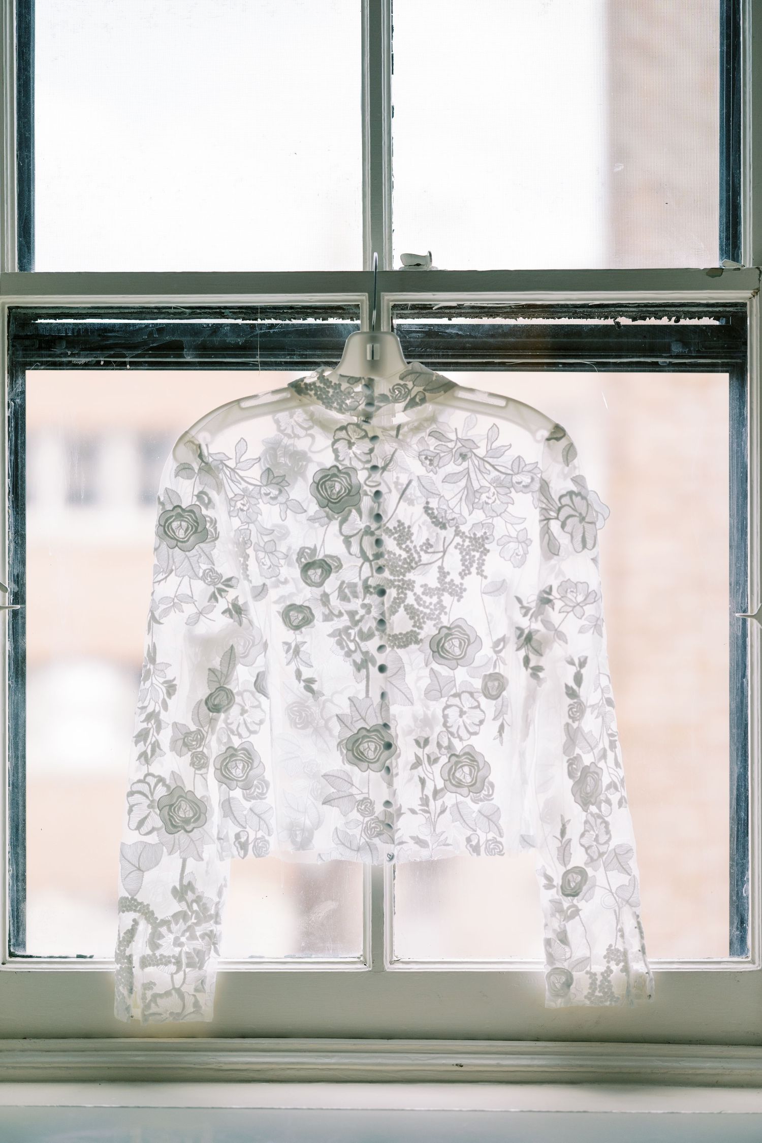 A delicate white lace jacket with floral patterns hangs in front of a sunlit window frame.
