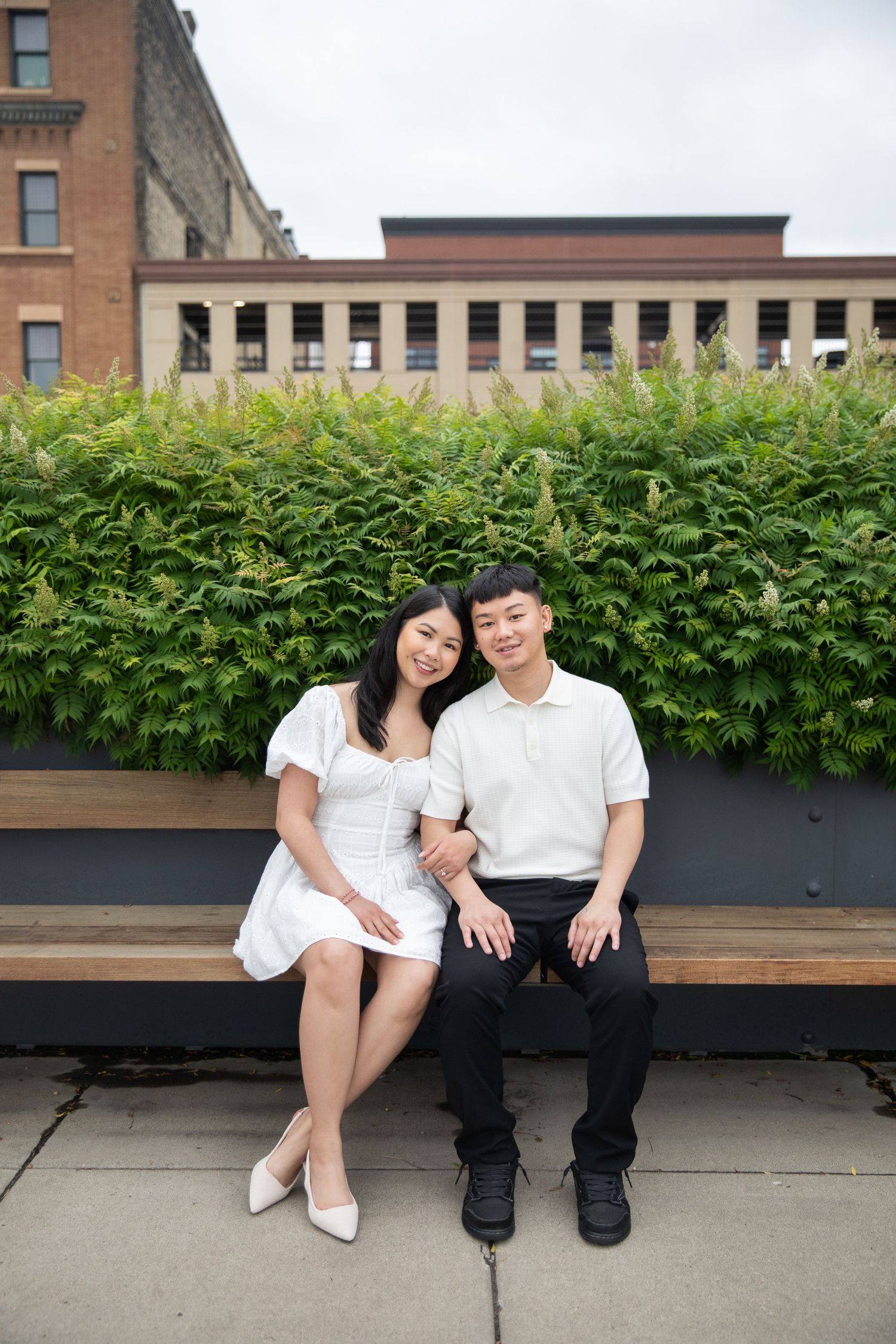 Two people in white shirts sit together on a wooden bench beside a green hedge planter in an urban setting.