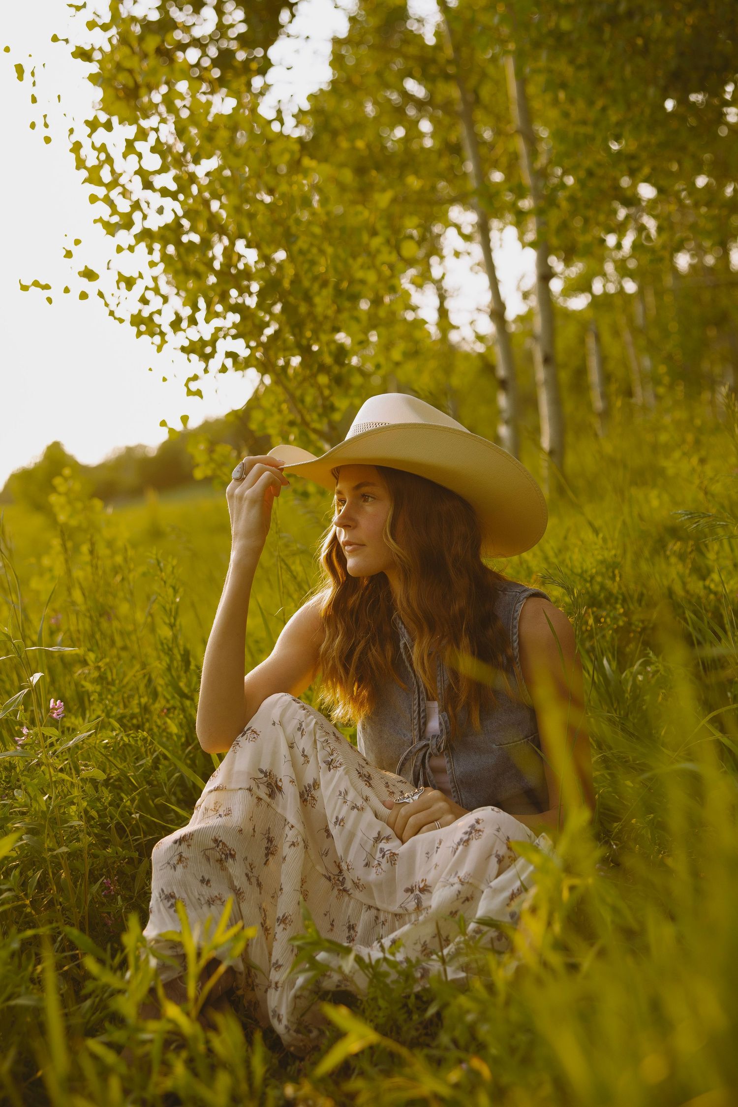Woman in white dress and cowboy hat sits in sunlit meadow surrounded by tall grass and birch trees.