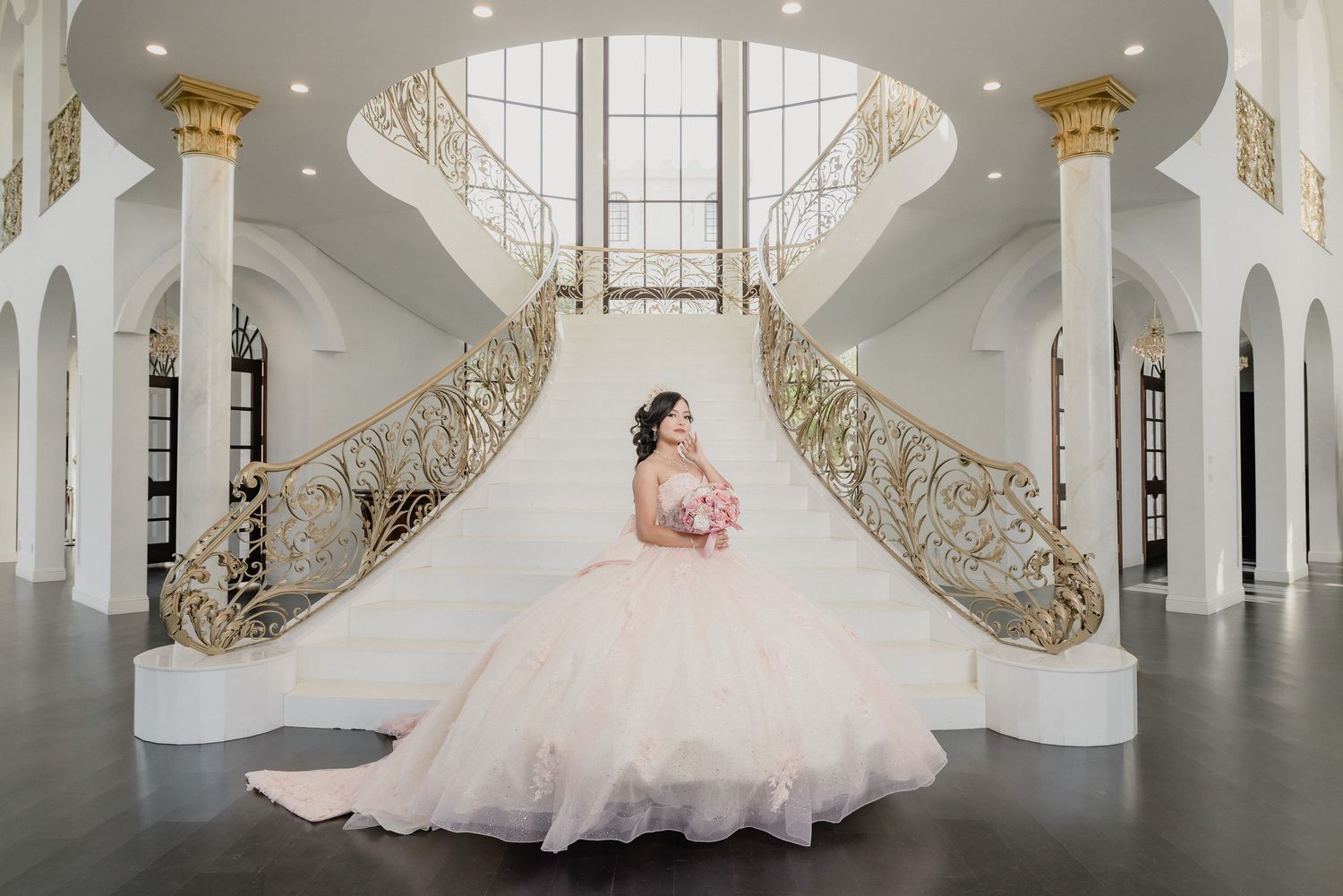 Elegant bride in flowing gown stands in grand white interior with ornate gold-accented spiral staircase and classical architecture.