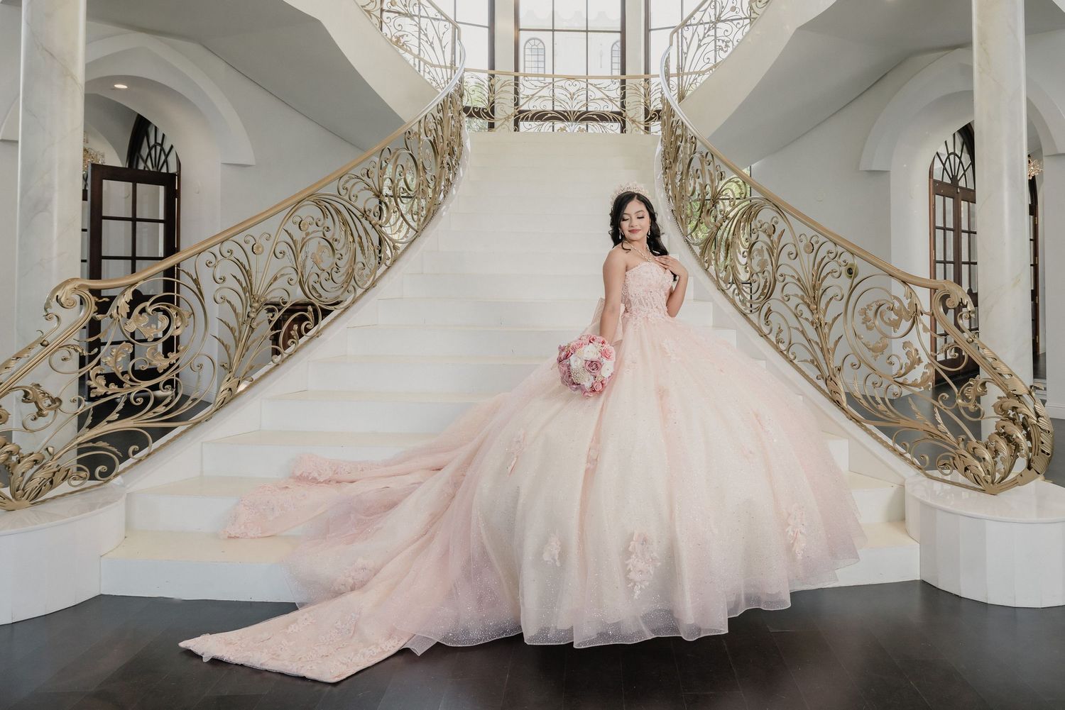 A light pink princess ballgown displayed in a grand white architectural space with decorative railings and windows.