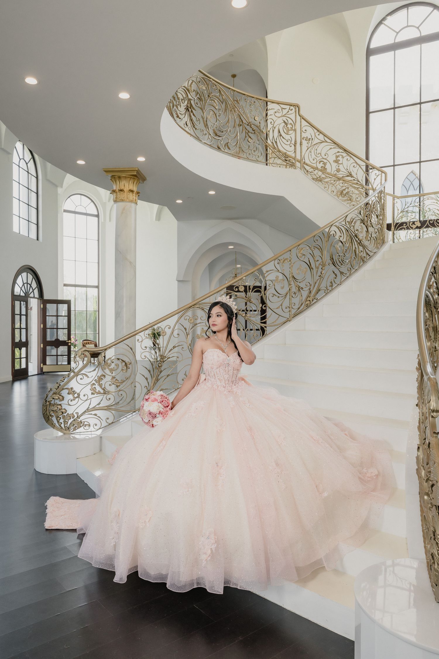 Elegant spiral staircase in white mansion interior with ornate gold railings and someone wearing a flowing pink ballgown.