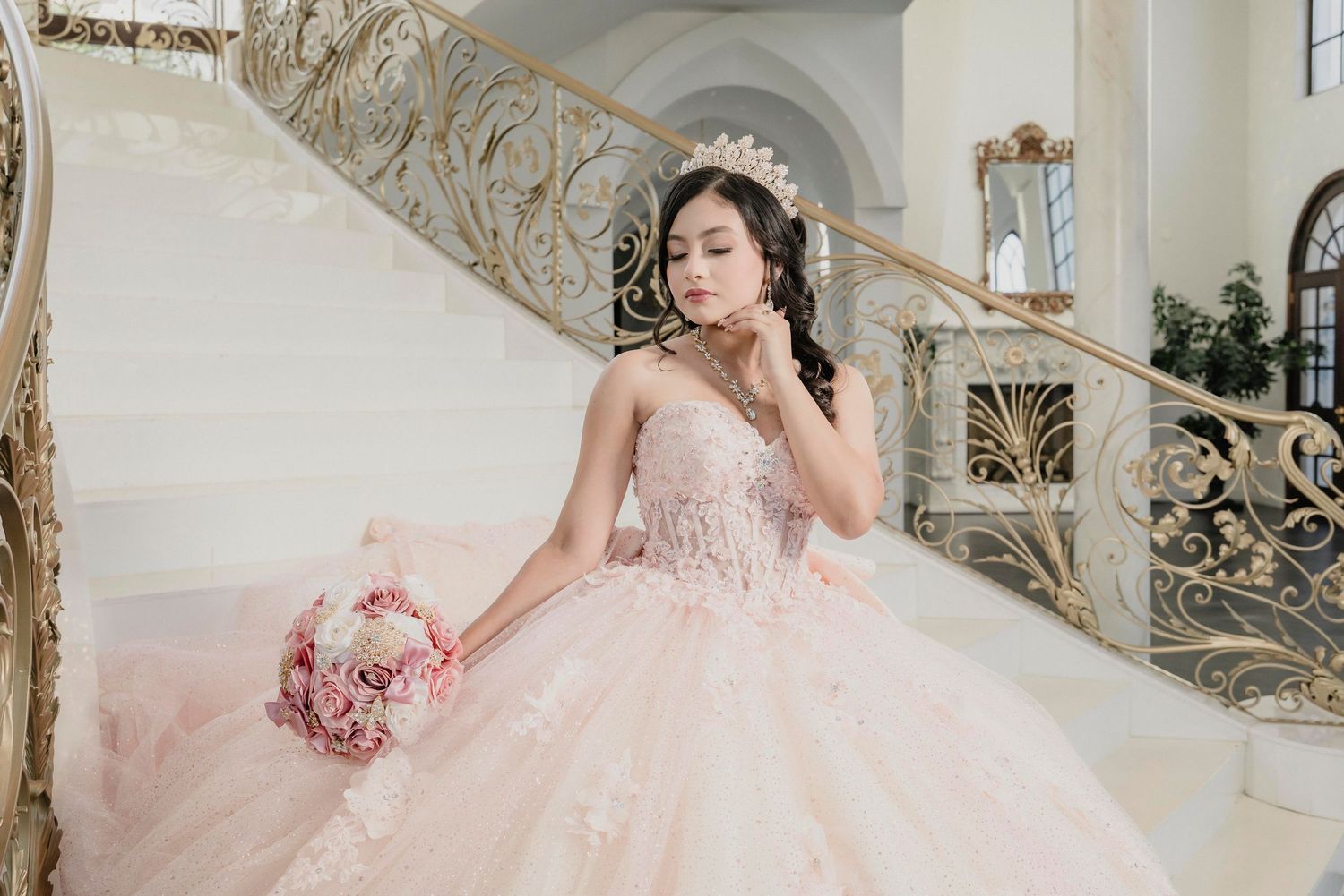 Elegant bride in intricate blush pink ballgown poses gracefully against ornate white and gold architectural backdrop.