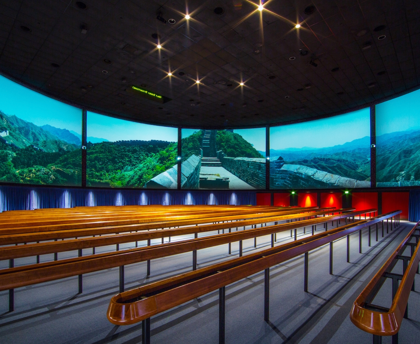 Modern theater with curved panoramic screen showing mountain scenery and tiered wooden bench seating.