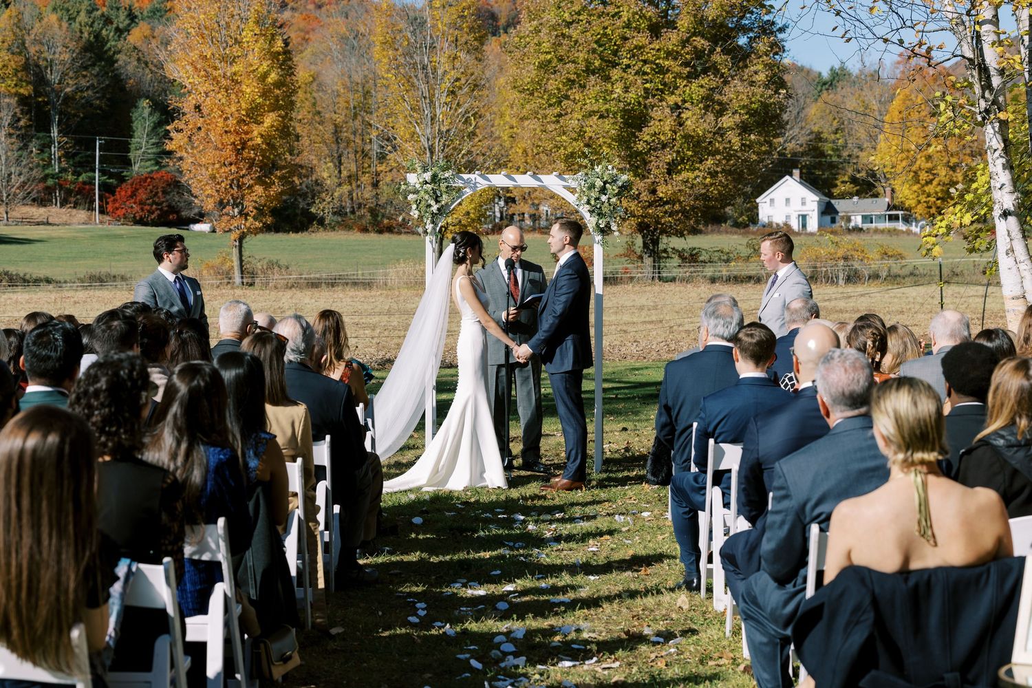 Outdoor autumn wedding ceremony taking place on grassy lawn surrounded by colorful fall foliage and white church in background.