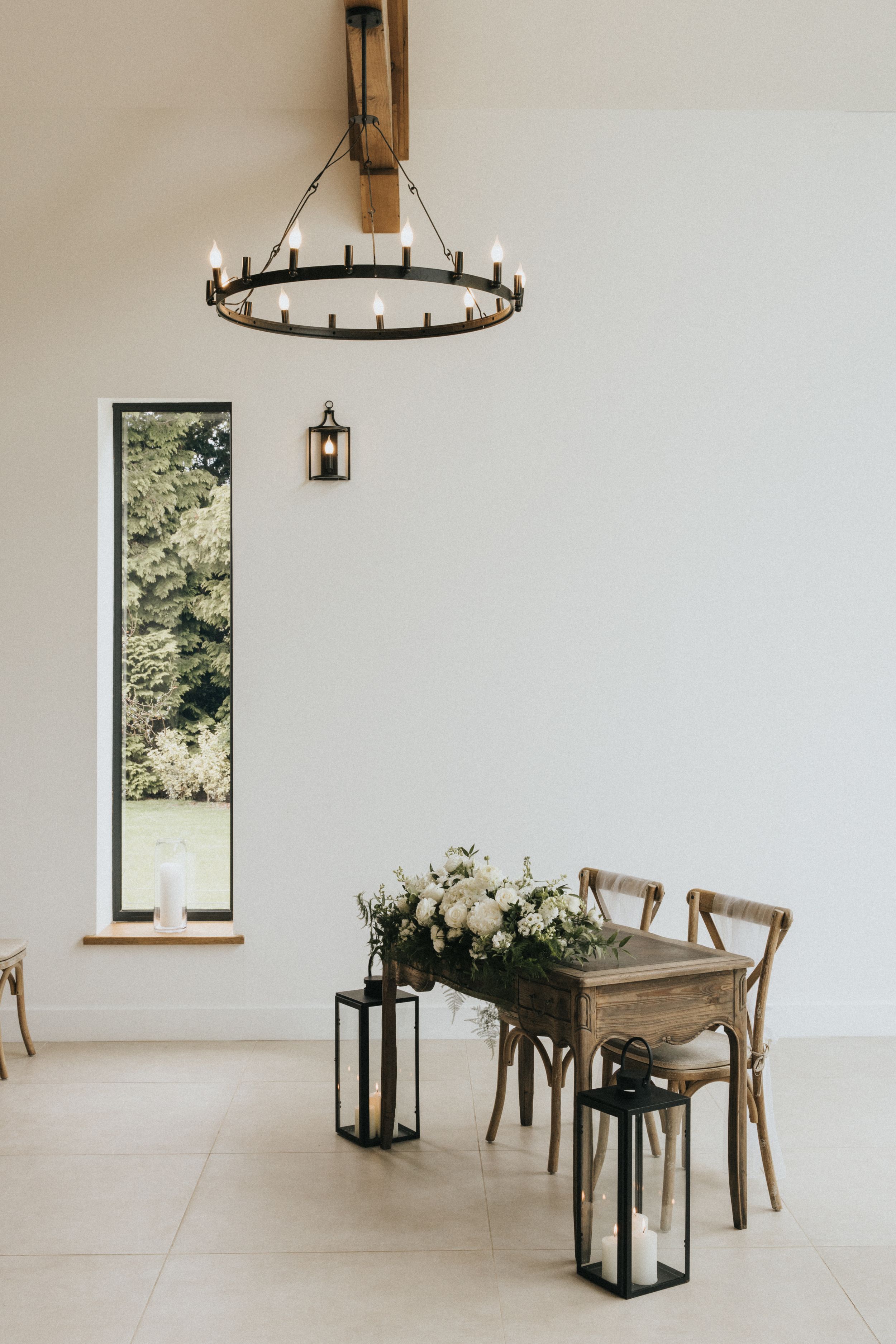 Modern minimalist dining space with circular iron chandelier, tall window, and rustic wooden chairs arranged around floral decor.