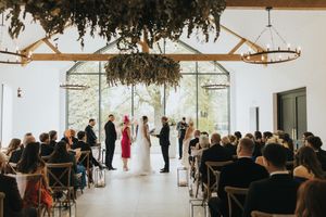 Wedding ceremony taking place in a modern white venue with large windows, hanging greenery and seated guests.