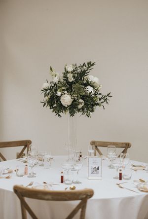 White tablescape with floral centerpiece, glassware, and rustic wooden chairs in a minimalist reception setting.