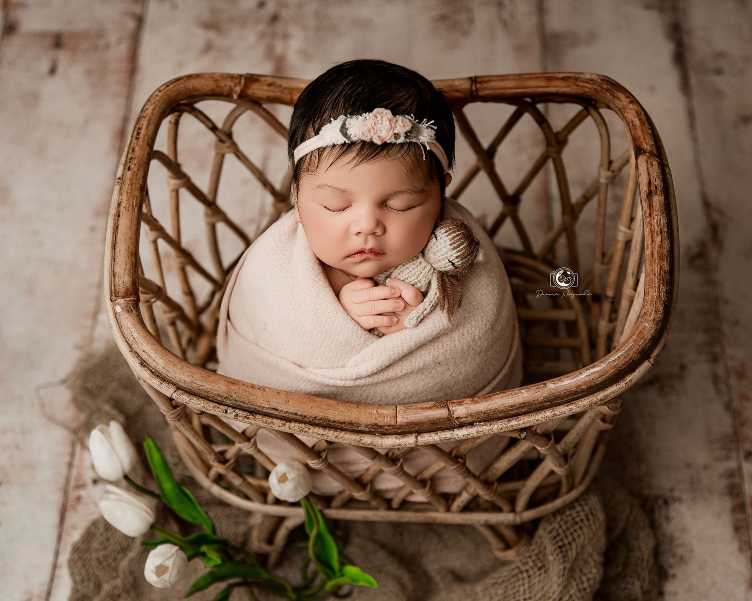 Newborn baby wrapped in beige cloth sleeps peacefully in woven rattan basket with delicate floral headband.
