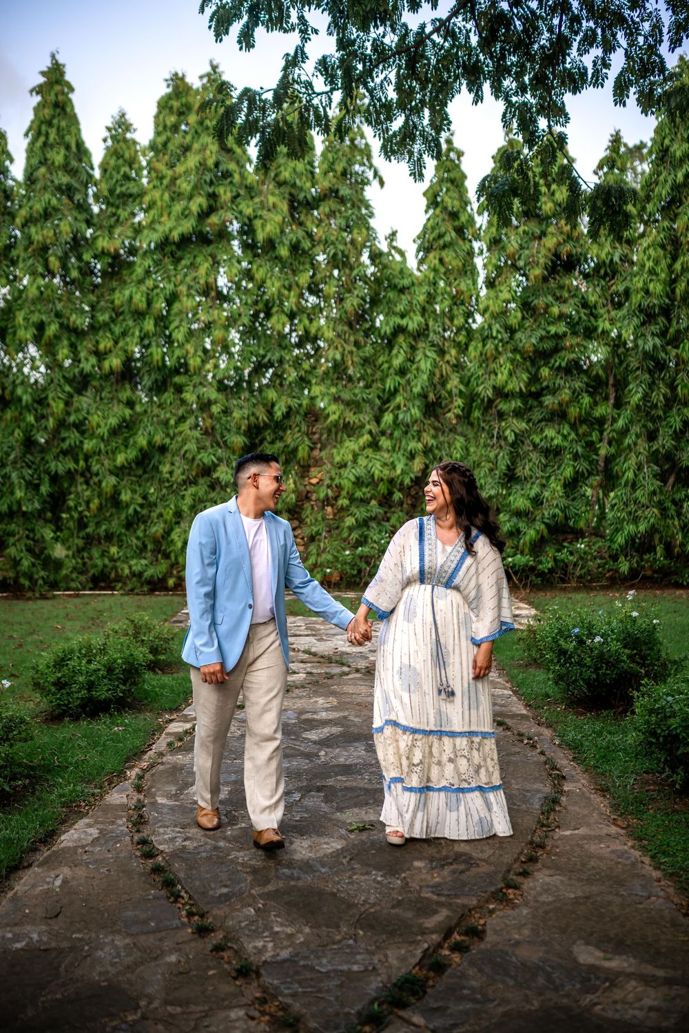 A couple walks hand in hand along a stone path surrounded by lush green trees and foliage in a garden setting.