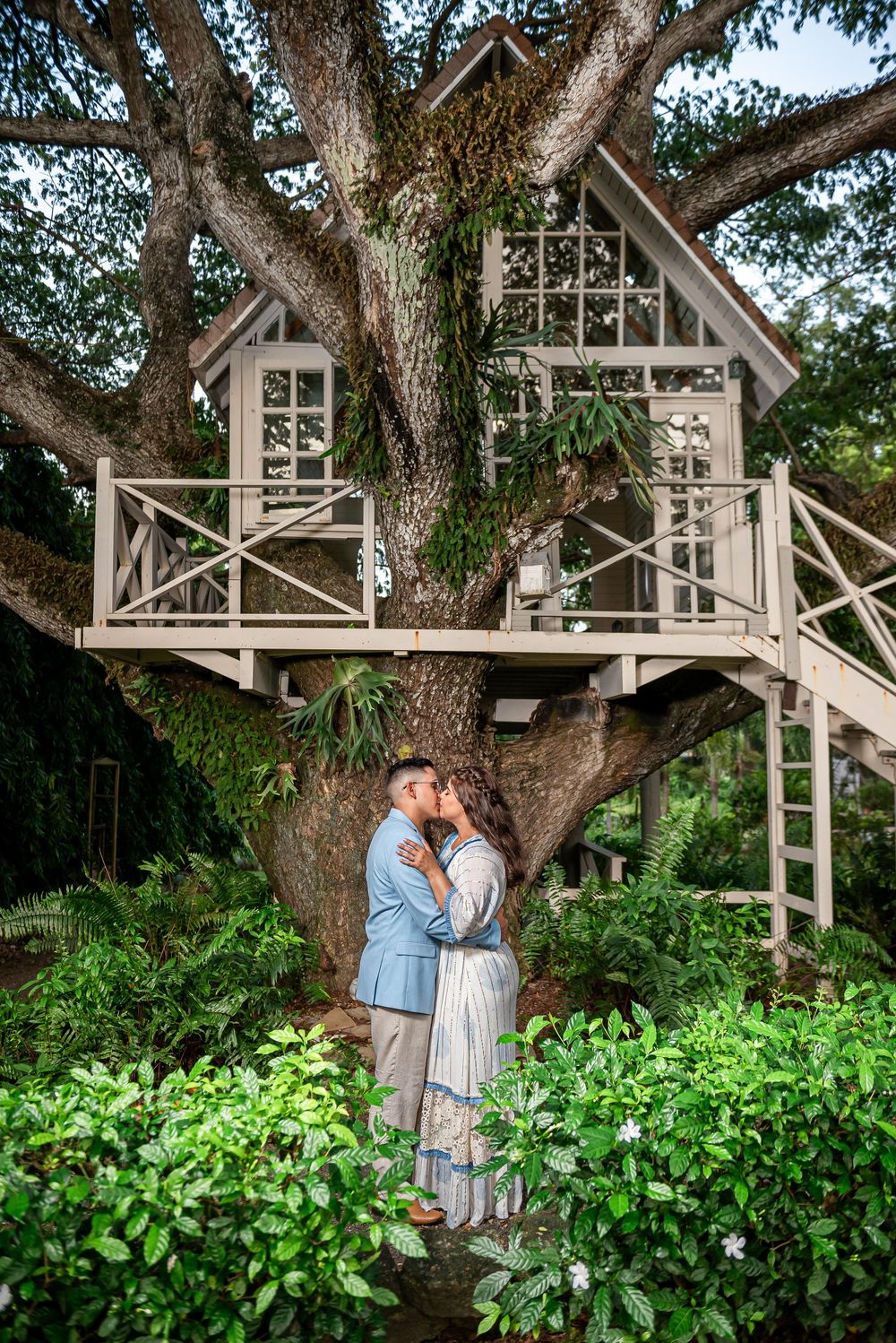 A charming white treehouse with glass windows nestled in a large tree surrounded by lush greenery.