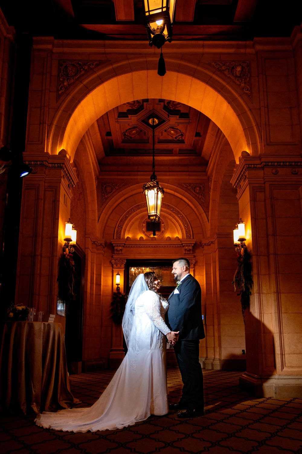 A romantic wedding couple stands beneath illuminated stone archways in an ornate historic building.