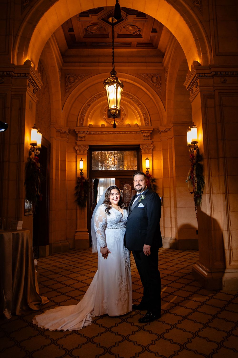 A newlywed couple poses under an elegant arched entrance illuminated by vintage lanterns at a historic venue.